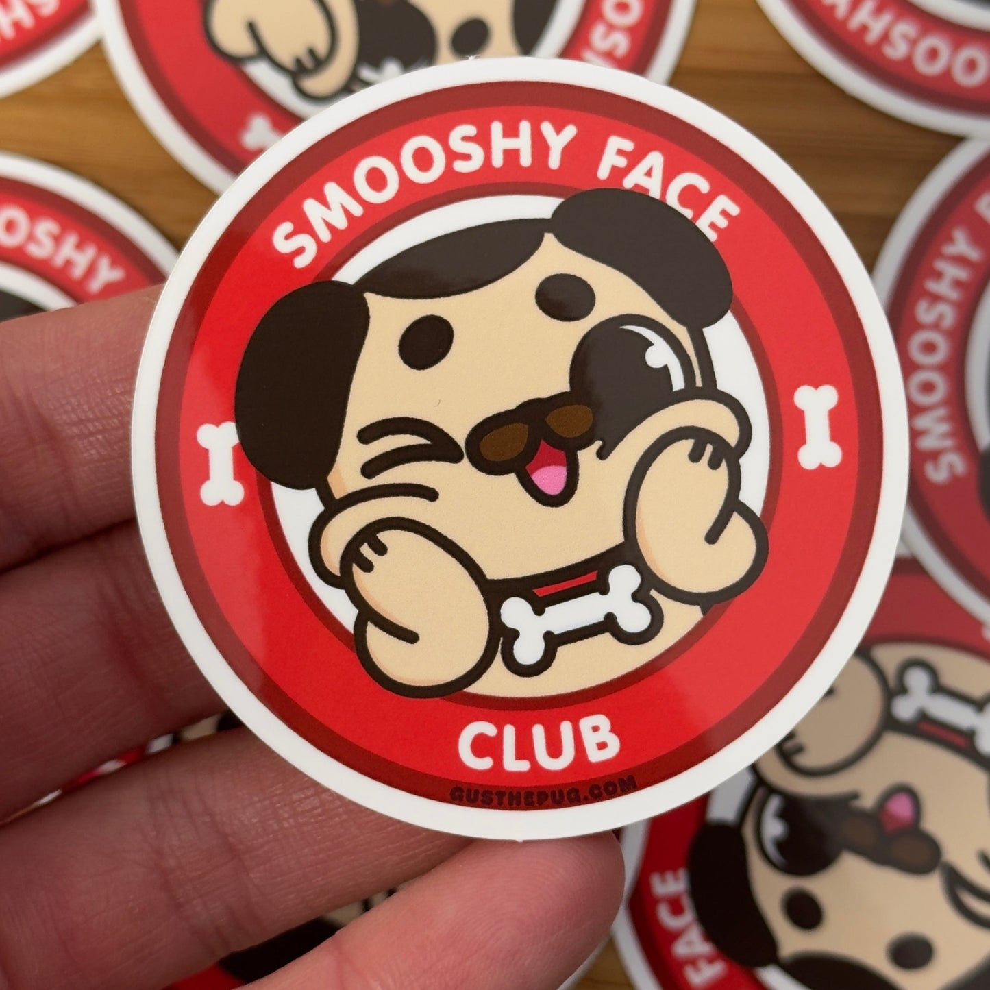 Sticker with 'Smooshy Face Club' design held by a hand, surrounded by more stickers on a wooden surface.